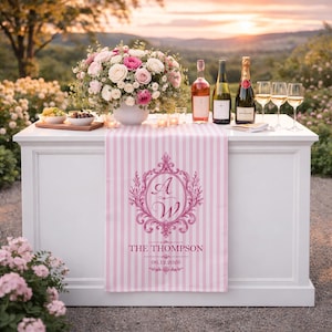 May include: A white bar table set for a celebration, with a pink and white striped table runner featuring a monogram and the name "The Thompson." The table is adorned with wine bottles, champagne, glasses, and a floral arrangement.