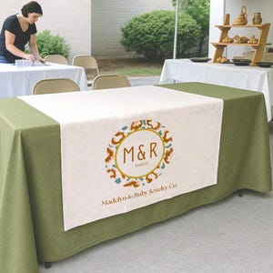 May include: A green tablecloth with a white table runner featuring a circular logo with the letters "M&R" and the words "Madelyn & Ruby Jewelry Co." A person is setting up a table in the background.