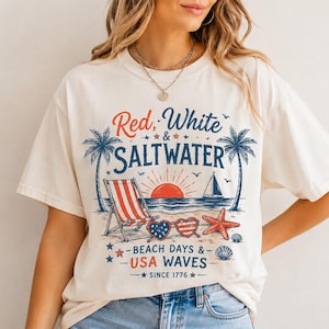 May include: A cream-colored short-sleeve t-shirt featuring a graphic print with the text Red, White & SALTWATER, BEACH DAYS & USA WAVES, and SINCE 1776. The design includes palm trees, a beach chair, a sailboat, heart-shaped sunglasses, and a starfish.
