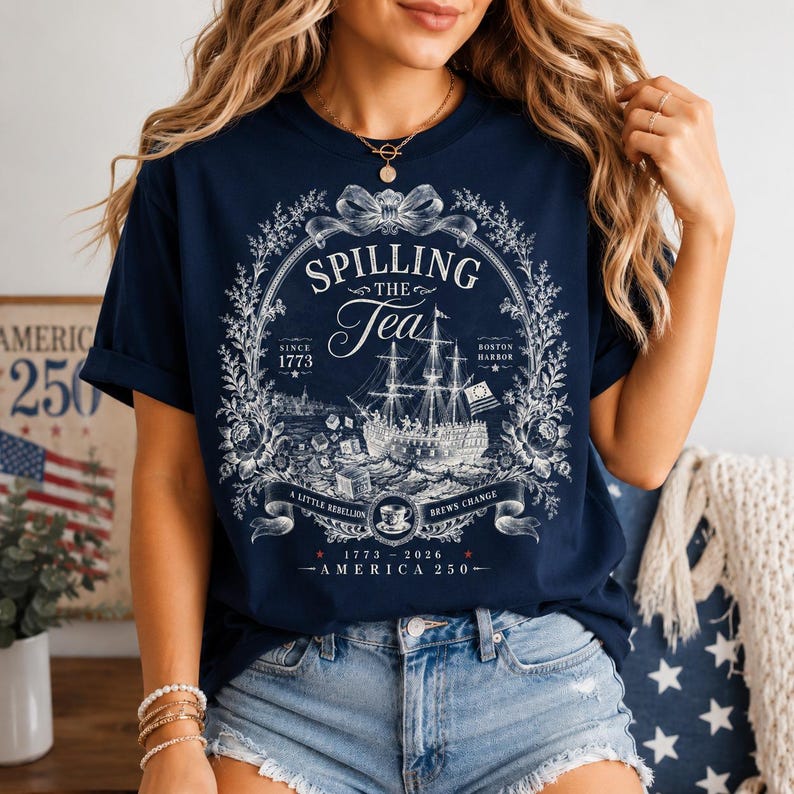 May include: A navy blue t-shirt featuring a white graphic print of a sailing ship, floral borders, and text that reads: Spilling The Tea, Since 1773, Boston Harbor, A Little Rebellion Brews Change, 1773-2026, and America 250.