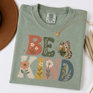 May include: A sage green short-sleeve t-shirt featuring the text BE KIND in large, colorful block letters. The letters are decorated with floral illustrations and a butterfly graphic. The shirt has a Comfort Colors brand label at the neck.