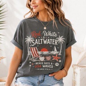 May include: A charcoal gray short-sleeve t-shirt featuring a graphic print with the text Red, White & SALTWATER, BEACH DAYS & USA WAVES, and SINCE 1776. The design includes palm trees, a beach chair, a sailboat, heart-shaped sunglasses, and a starfish.