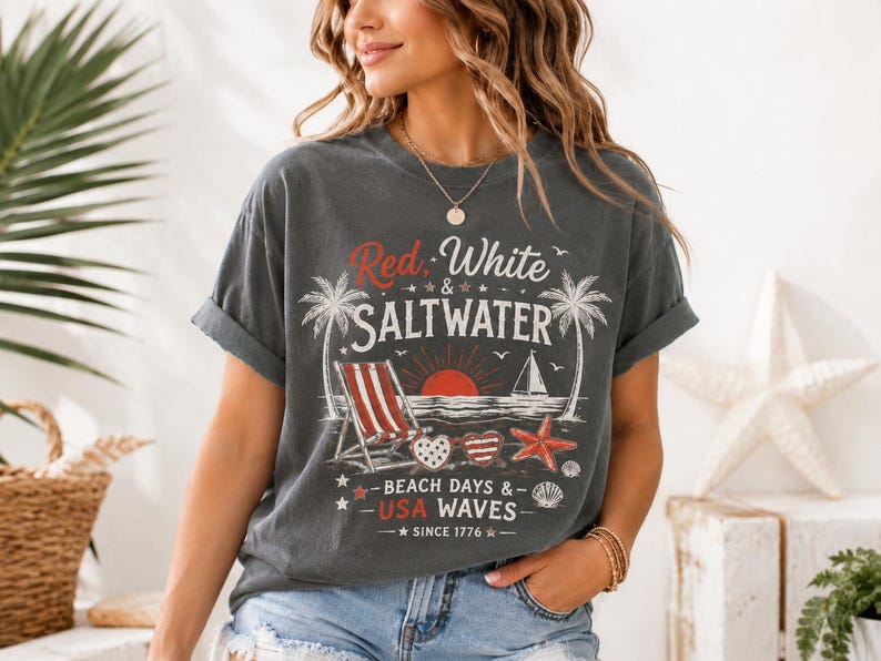 May include: A charcoal gray short-sleeve t-shirt featuring a graphic print with the text Red, White & SALTWATER, BEACH DAYS & USA WAVES, and SINCE 1776. The design includes palm trees, a beach chair, a sailboat, heart-shaped sunglasses, and a starfish.