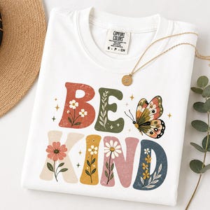 May include: A white short-sleeve t-shirt with a distressed graphic print that reads BE KIND in large, multi-colored block letters. The letters are decorated with floral illustrations and a butterfly. The shirt has a Comfort Colors brand label.