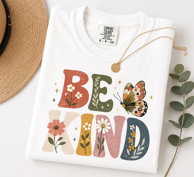 May include: A white short-sleeve t-shirt with a distressed graphic print that reads BE KIND in large, multi-colored block letters. The letters are decorated with floral illustrations and a butterfly. The shirt has a Comfort Colors brand label.