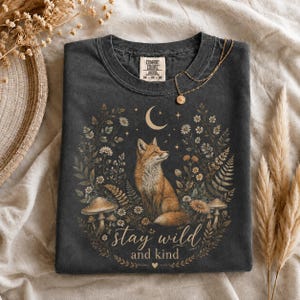 May include: A charcoal gray t-shirt featuring a graphic of a fox surrounded by flowers, ferns, mushrooms, and a crescent moon. The text "stay wild and kind" is printed below the fox. The shirt has a Comfort Colors brand label at the neckline.