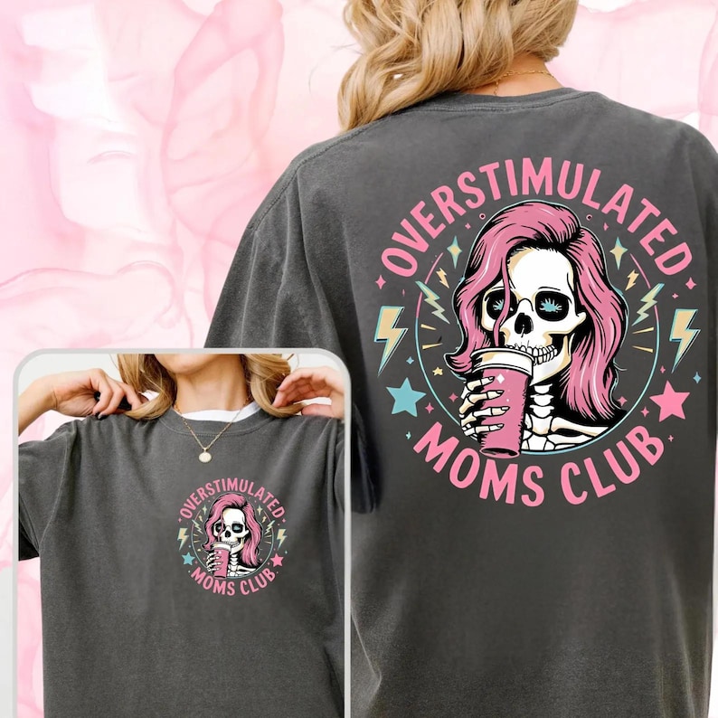 Pu&ograve; includere: Una maglietta grigia scura con una grafica di teschio rosa e nero. Il teschio indossa una parrucca rosa e tiene una tazza rosa. Il testo "Overstimulated Moms Club" &egrave; scritto in un cerchio attorno al teschio.