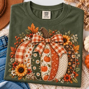 Puede incluir: Una camiseta verde oliva con un gran gráfico central de una calabaza. La calabaza está compuesta por varios segmentos estampados que incluyen cuadros, flores y texturas de punto, rematada con un gran lazo de cuadros naranja y blanco.