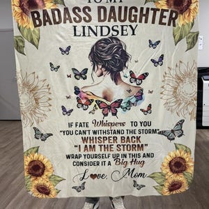 Op de afbeelding: Een crèmekleurige fleece deken met zonnebloemen in de hoeken, vlinders en een illustratie van een persoon van achteren. De tekst luidt: TO MY BADASS DAUGHTER LINDSEY, IF FATE WHISPERS TO YOU YOU CAN'T WITHSTAND THE STORM WHISPER BACK I AM THE STORM WRAP YOURSELF UP IN THIS AND CONSIDER IT A BIG HUG Love, Mom.