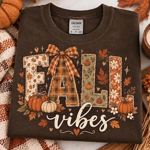 May include: A dark brown t-shirt featuring the text FALL in large block letters filled with floral, plaid, and pumpkin patterns, topped with a plaid bow. The word vibes is written below in white script, surrounded by autumn leaves and pumpkins.