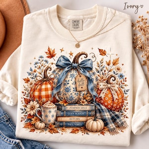 May include: An ivory crewneck t-shirt featuring a graphic of three pumpkins, a mug of coffee, and a stack of books. The central pumpkin has a blue bow and a tag reading hello fall. The books are labeled pumpkin spice, cozy season, autumn, fall vibes.