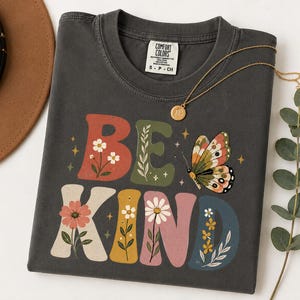 May include: A charcoal gray short-sleeve t-shirt featuring the text BE KIND in large, colorful, block-style letters. The letters are decorated with floral illustrations and a butterfly graphic. The shirt has a Comfort Colors brand label.