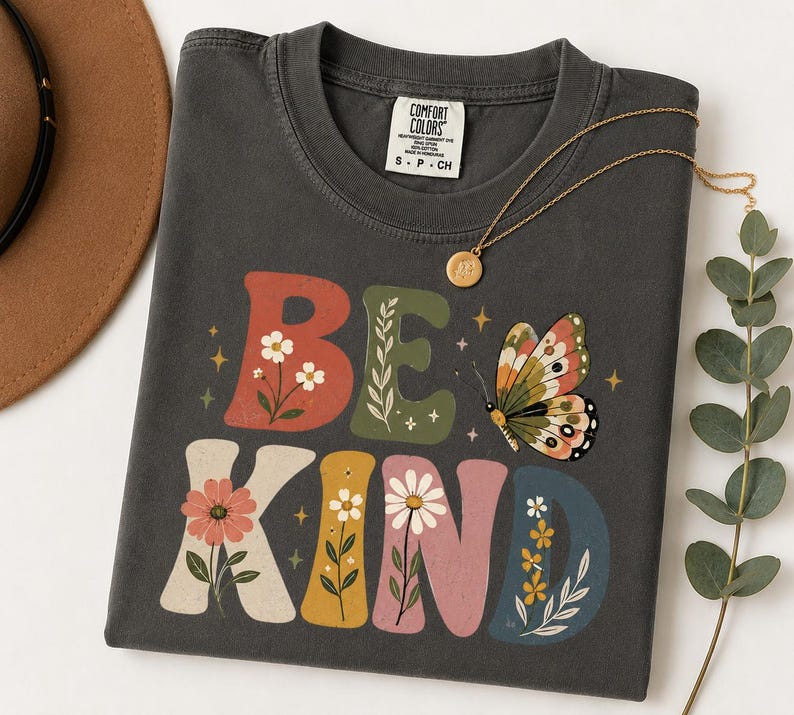 May include: A charcoal gray short-sleeve t-shirt featuring the text BE KIND in large, colorful, block-style letters. The letters are decorated with floral illustrations and a butterfly graphic. The shirt has a Comfort Colors brand label.