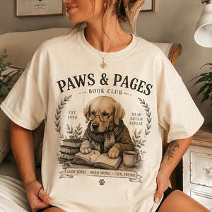 Op de afbeelding: Een crèmekleurig t-shirt met een afbeelding van een golden retriever puppy met een bril en een trui aan, die een boek leest. De tekst luidt PAWS & PAGES BOOK CLUB, EST. 1998, READ DREAM REPEAT en GOOD BOOKS WARM DRINKS LOYAL FRIENDS.
