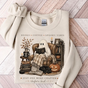 Könnte beinhalten: Ein cremefarbenes Sweatshirt mit Rundhalsausschnitt und einer Grafik eines Geistes, der in einem Sessel ein Buch liest. Der Text lautet "BOOKS • COFFEE • SPOOKY VIBES" und "JUST ONE MORE CHAPTER before bed". Die Illustration zeigt eine schwarze Katze und Kürbisse.