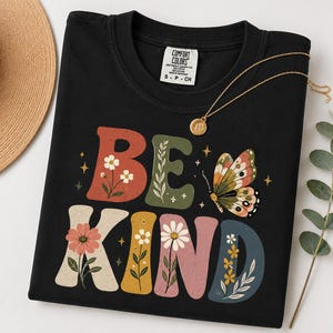 May include: A black t-shirt featuring the text BE KIND in large, colorful, block-style letters decorated with floral illustrations and small stars. A butterfly graphic is positioned next to the word BE, and the shirt has a Comfort Colors brand label.