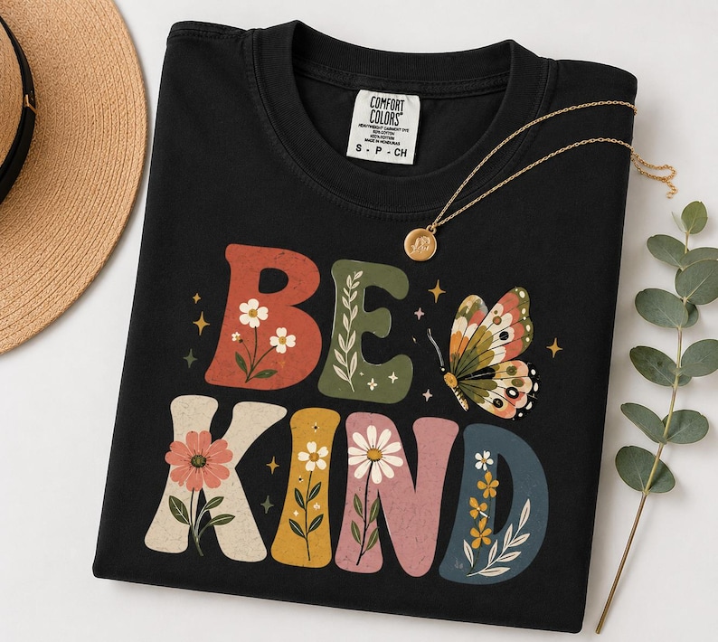May include: A black t-shirt featuring the text BE KIND in large, colorful, block-style letters decorated with floral illustrations and small stars. A butterfly graphic is positioned next to the word BE, and the shirt has a Comfort Colors brand label.