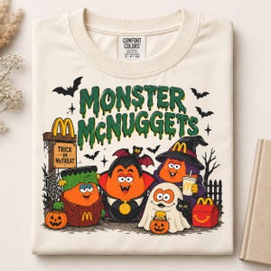 May include: A cream-colored t-shirt featuring a graphic with the text MONSTER MCNUGGETS in green. The design shows four orange nugget characters dressed as a Frankenstein monster, a vampire, a witch, and a ghost, with a McDonald's sign.