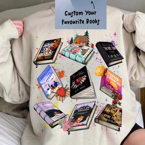 Puede incluir: Sudadera beige con ilustraciones de varios libros con títulos como "Incent", "Between Friends Lovers" y "Seven Summers". El texto "Custom Your Favourite Books" está en la parte superior. El diseño incluye acentos coloridos y elementos florales.