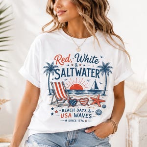 May include: A white t-shirt featuring a graphic print with the text Red, White & SALTWATER and BEACH DAYS & USA WAVES SINCE 1776. The design includes palm trees, a beach chair, a sailboat, a sun, heart-shaped sunglasses, a starfish, and seashells.