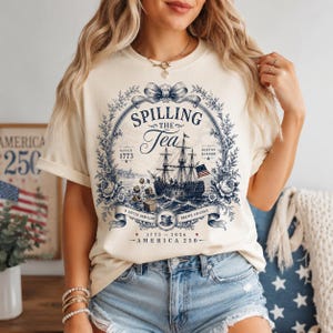 May include: A cream-colored t-shirt featuring a dark blue graphic of a historic sailing ship, a floral wreath, and the text "SPILLING THE TEA", "SINCE 1773", "BOSTON HARBOR", "A LITTLE REBELLION BREWS CHANGE", and "1773-2026 AMERICA 250".