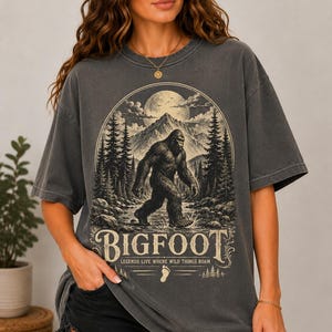 May include: A charcoal gray short-sleeve t-shirt featuring a graphic print of Bigfoot walking through a forest with mountains and a full moon in the background. The text reads BIGFOOT and LEGENDS LIVE WHERE WILD THINGS ROAM.