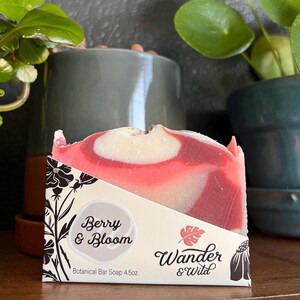 Könnte beinhalten: Ein botanisches Stück Seife mit einem Beeren- und Blüten-Design. Die Seife hat einen Wirbel aus roten und weißen Farben. Die Verpackung trägt die Aufschrift "Berry & Bloom" und "Wander & Wild". Die Seife wiegt 128 Gramm.