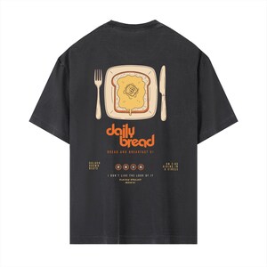 Puede incluir: Una camiseta gris carbón de manga corta con un gráfico de una rebanada de pan tostado con mantequilla en un plato, junto a un tenedor y un cuchillo. El texto dice daily bread, BREAD AND BREAKFAST VI, GOLDEN BROWN BEATS, DAILY BREAD MUSIC.
