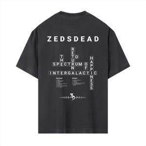 May include: Dark gray t-shirt with the text "ZEDSDEAD" at the top. Below, a crossword puzzle design spells out "Return to the Spectrum of Happiness" and "Intergalactic". The shirt also features a stylized heartbeat graphic.
