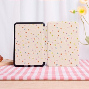 Pode incluir: Uma capa de tablet bege com uma borda preta, aberta para revelar um interior estampado. A capa e o interior apresentam um padr&atilde;o repetitivo de estrelas coloridas em tons de vermelho, azul, verde e roxo. A capa est&aacute; sobre uma superf&iacute;cie xadrez rosa e branca.