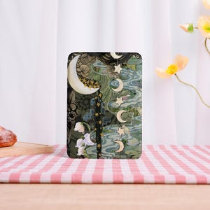 May include: A rectangular e-reader case featuring a dark green background with a large white crescent moon, a vertical row of smaller moon phases and stars, and white lily flowers. The design includes intricate line art and flowing water patterns.