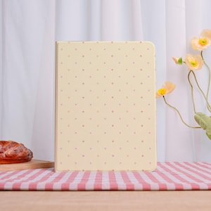 Peut inclure: Une housse de tablette jaune clair avec un motif à pois roses. L'étui est rectangulaire et se dresse sur une surface en bois. L'arrière-plan comprend un rideau blanc, une nappe à carreaux roses et blancs et des fleurs décoratives.