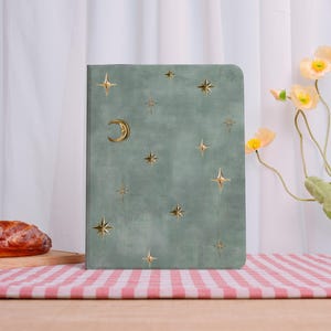May include: A sage green journal with a celestial design. Gold-colored stars and a crescent moon adorn the cover. The journal is rectangular and has a textured appearance. The background includes flowers and a red and white checkered tablecloth.