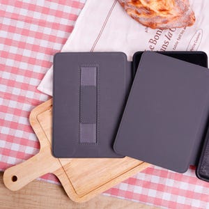 Pode incluir: Uma capa cinza para tablet com uma al&ccedil;a el&aacute;stica para a m&atilde;o est&aacute; aberta em uma t&aacute;bua de cortar de madeira. A capa est&aacute; em uma toalha de mesa xadrez vermelha e branca, com um peda&ccedil;o de p&atilde;o e um pano branco no fundo.