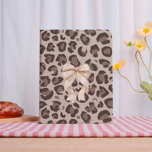 May include: A rectangular journal with a beige leopard print cover and a cream-colored bow. The journal is closed and sits on a red and white checkered tablecloth. The background includes a white curtain and yellow flowers.