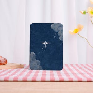 May include: A rectangular phone case featuring an aerial view of a white airplane flying through a dark blue sky with fluffy gray clouds. The case is set against a red and white checkered tablecloth.