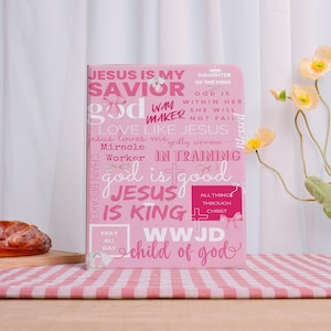 Peut inclure: Journal rose avec des inscriptions religieuses, dont "Jésus est mon Sauveur" et "Enfant de Dieu". La couverture présente diverses phrases et mots en blanc et rose, avec des croix et des nœuds. Le journal est sur une nappe à carreaux roses et blancs.
