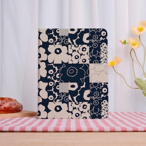 May include: A hardcover journal with a navy blue and cream floral pattern. The design features a patchwork of stylized flowers in various sizes and shapes. The journal is rectangular and stands upright on a wooden surface.