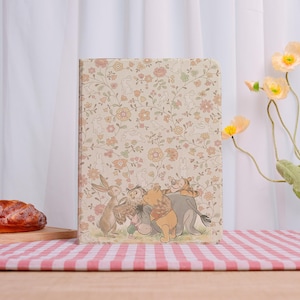 May include: A rectangular journal with a floral and rabbit pattern. The cover features a scene with Winnie the Pooh and friends. The background is a beige color with orange and pink flowers and white rabbits.