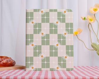 May include: A hardcover notebook with a daisy and checkerboard pattern. The cover features a repeating design of white daisies with yellow centers set against a green and cream checkerboard background. The notebook is displayed on a pink and white checkered tablecloth.
