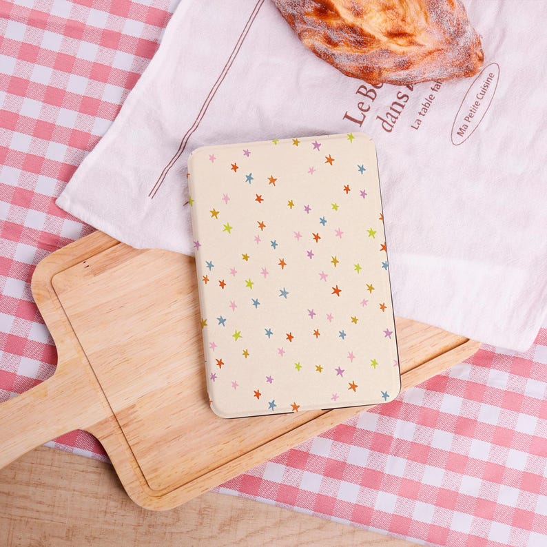 Pode incluir: Um caderno creme com um padr&atilde;o de estrelas coloridas. O caderno est&aacute; sobre uma t&aacute;bua de cortar de madeira, com um pano branco e um p&atilde;o no fundo. O fundo tamb&eacute;m inclui uma toalha de mesa xadrez vermelha e branca.