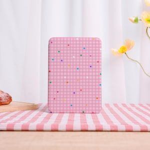 May include: A pink rectangular case featuring a gingham design and multi-coloured star shapes. The case is placed on a pink and white checked surface. The background includes a white curtain and some flowers.
