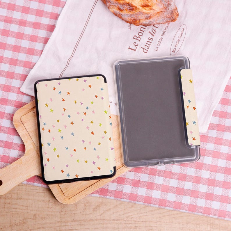 Pode incluir: Uma capa de tablet aberta com uma capa creme adornada com estrelas coloridas. A capa est&aacute; em uma t&aacute;bua de corte de madeira, ao lado de uma capa de pl&aacute;stico transparente. O fundo inclui um pano branco com texto em franc&ecirc;s e um padr&atilde;o xadrez rosa e branco.