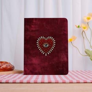 May include: A rectangular notebook with a deep burgundy, textured cover. Centered on the front is a red heart illustration featuring a central blue eye, surrounded by radiating gold lines, small white stars, and a border of white pearls.