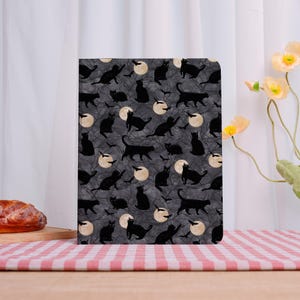 May include: A rectangular notebook featuring a pattern of black cats, bats, and full moons on a dark grey backdrop. The design creates a Halloween or spooky aesthetic.