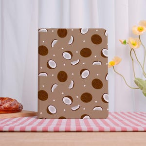 May include: A brown notebook with a repeating pattern of coconuts and white dots. The coconuts are whole and sliced, with a white interior. The notebook is on a pink and white checkered surface.