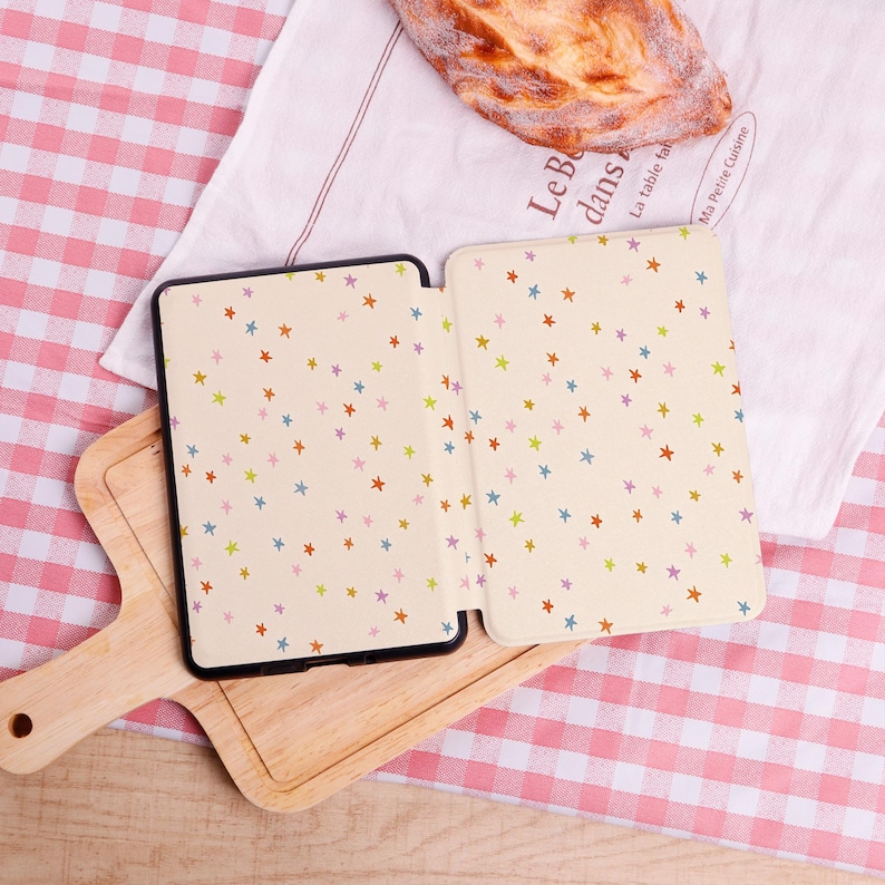 Pode incluir: Uma capa de tablet aberta com fundo creme e um padr&atilde;o de estrelas coloridas. A capa est&aacute; sobre uma t&aacute;bua de cortar de madeira, com uma toalha de mesa xadrez vermelha e branca ao fundo. Um p&atilde;o descansa sobre um pano branco.