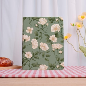 May include: A notebook with a floral pattern of cream-colored roses and green leaves on a sage green background. The notebook is rectangular and stands upright on a wooden surface with a red and white checkered tablecloth.