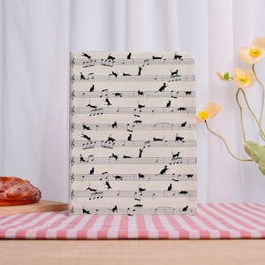 May include: A notebook featuring a musical staff design with black cat silhouettes in various poses. The notebook has a cream-colored background and is displayed on a wooden surface with a red and white checkered tablecloth.
