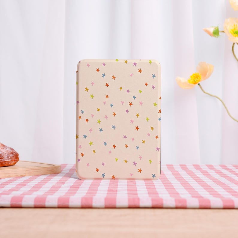 Pode incluir: Uma lata retangular bege com um padr&atilde;o repetido de estrelas coloridas. As estrelas s&atilde;o em tons de vermelho, laranja, azul, verde e roxo. A lata est&aacute; sobre uma superf&iacute;cie xadrez rosa e branca.